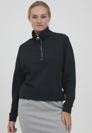 Sweat-shirt zippé noir avec un col haut, présentant un design en chevrons et des poignets côtelés. Tissu doux, coupe courte. Jupe grise visible en dessous.