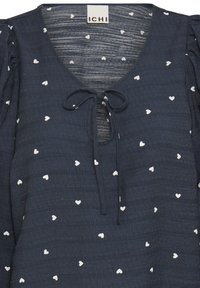 Blusa azul marino con mangas cortas abullonadas, escote en V y patrones blancos en forma de corazón. Presenta un cierre con lazo en el cuello y una tela texturizada.