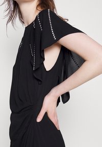 Black dress with ruffled sleeves, adorned with silver studs along the shoulder. Soft, draped fabric with a twisted detail at the waist.