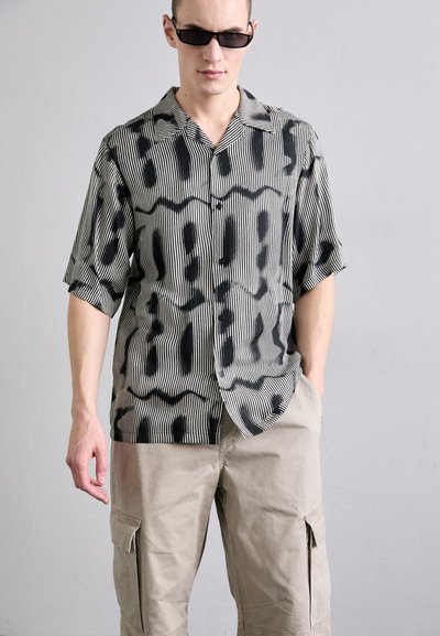 Chemise à manches courtes à rayures noires et blanches avec des motifs abstraits, faite en tissu léger, associée à un pantalon cargo beige.