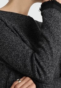 Žena oblečená v texturovaném tmavě šedém topu s dlouhým rukávem a černými detaily, spočívající bradou na ruce, s viditelným stříbrným prstenem na prstu.