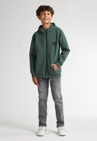 Zelená mikina s kapucí a zipem, přední kapsy a černé logo na hrudi, spárovaná se světle šedými džíny a bílými teniskami. Hladká textura.