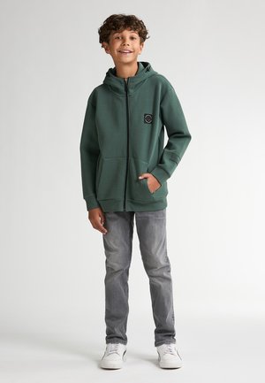 Sweat à capuche vert avec une fermeture éclair, poches avant, et un logo noir sur la poitrine, associé à un jean gris clair et des baskets blanches. Texture lisse.