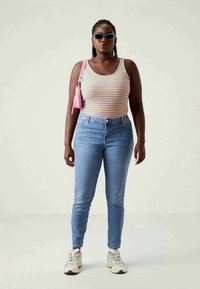 Mulher vestindo uma camisola de tiras, jeans azul claro, ténis, óculos de sol e a carregar uma pequena mala de ombro rosa, de pé contra um fundo simples.