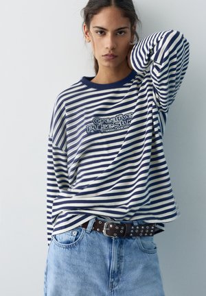 Giovane donna con capelli lunghi e scuri che indossa una maglia a maniche lunghe a righe blu navy e bianche con scritte, jeans azzurri e una cintura nera con occhielli metallici.