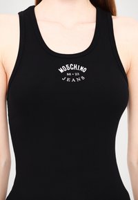 Melna ribota bez piedurknēm topa dizains ar apaļu dekoltē un platajām siksnām. Priekšpusē attēlots balts logo teksts "MOSCHINO 86 23 JEANS."