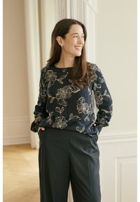 Šypsosi moteris su ilgais rudais plaukais, stovinti su rankomis kišenėse, dėvinti tamsią gėlėtą palaidinę ir juodus kelnes šviesiame kambaryje.