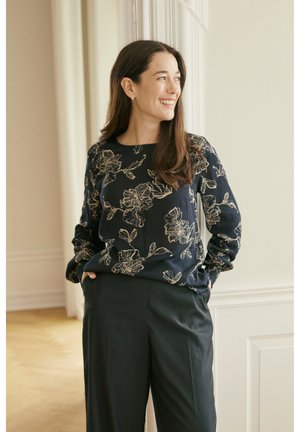 Smaidoša sieviete ar gariem brūniem matiem stāv ar rokām kabatās, valkājot tumšu ziedu raksta blūzi un melnas bikses gaišā istabā.