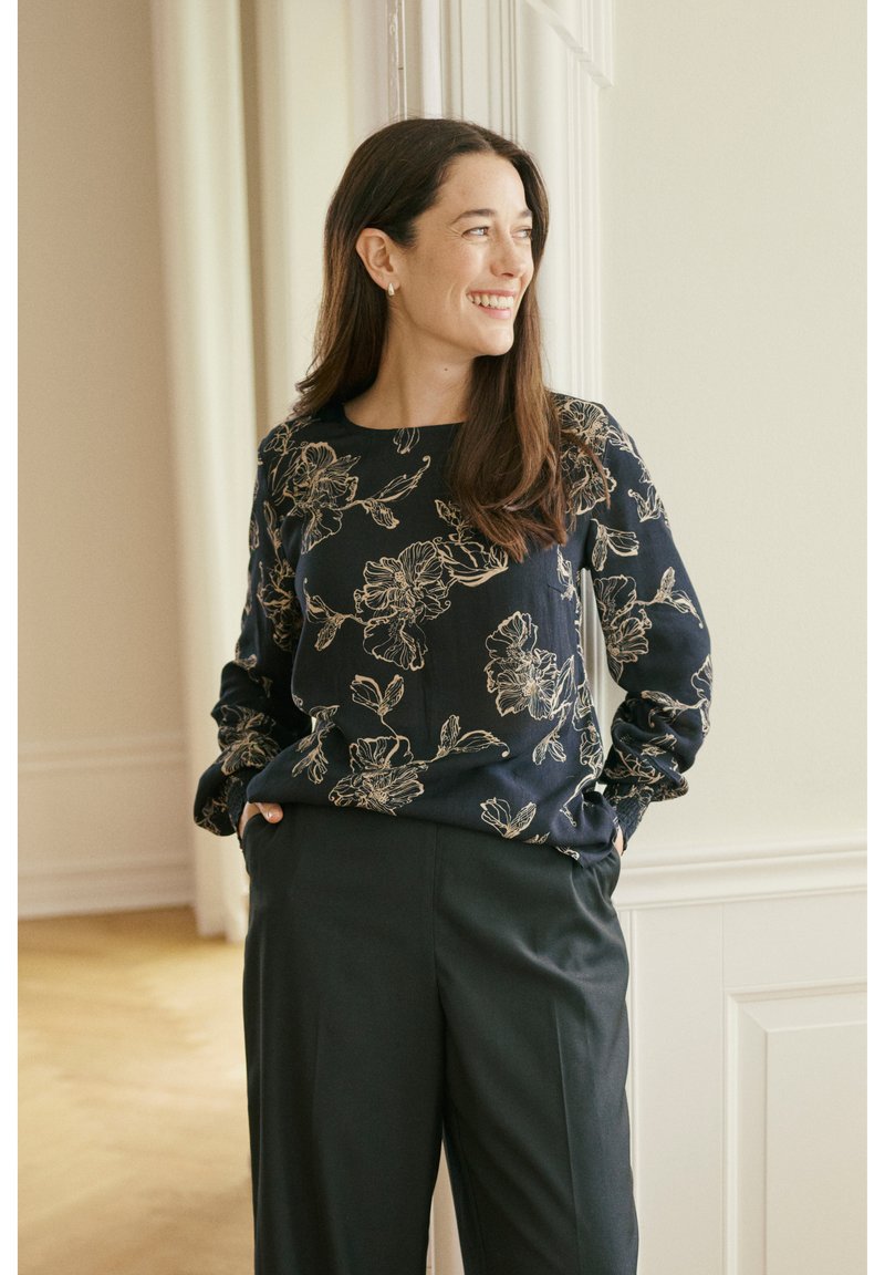 Šypsosi moteris su ilgais rudais plaukais, stovinti su rankomis kišenėse, dėvinti tamsią gėlėtą palaidinę ir juodus kelnes šviesiame kambaryje.