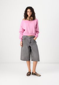 Felpa rosa scollata sulle spalle, culotte in denim grigio e décolleté con stampa animale. La modella sta in piedi con le mani nelle tasche su uno sfondo bianco.