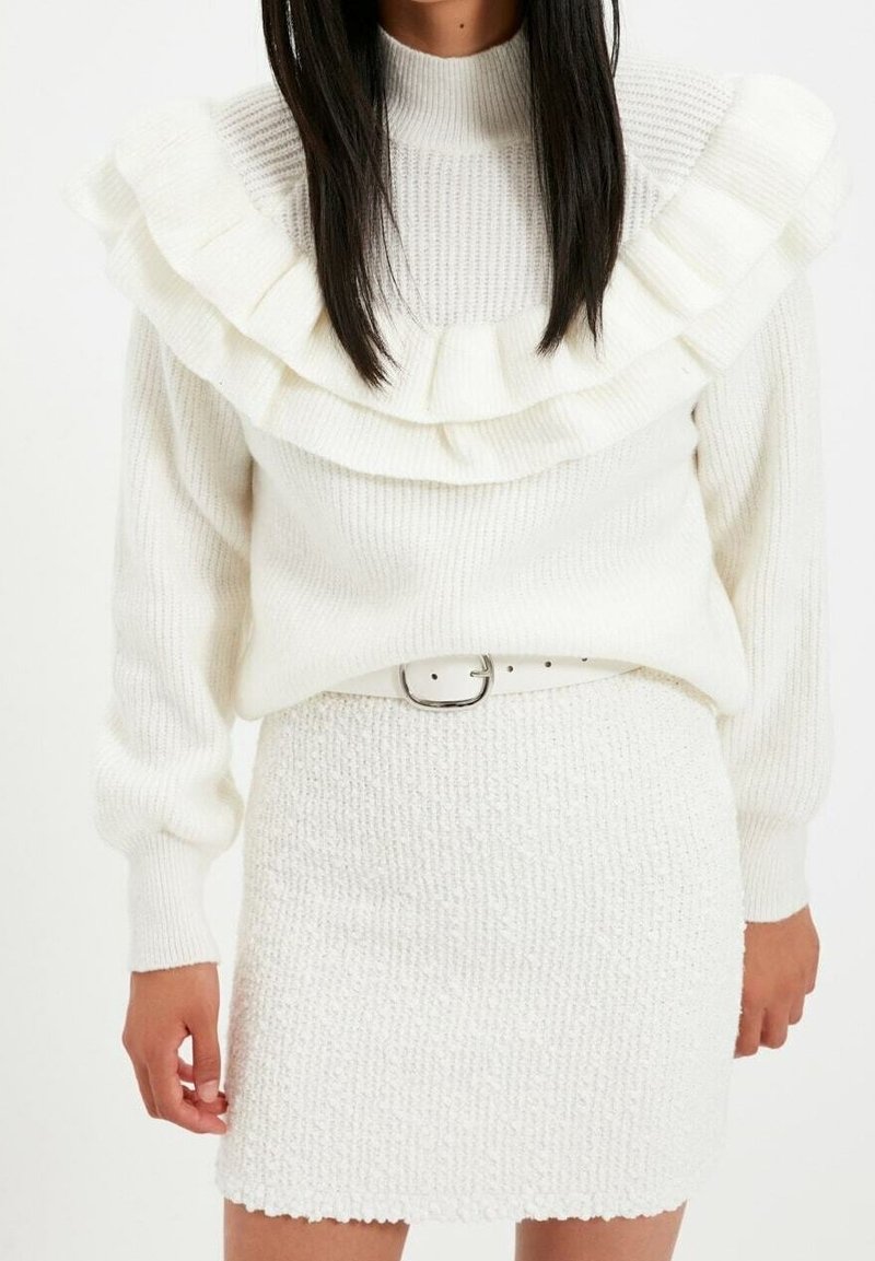 Femme portant un pull en maille texturée blanche avec des détails à volants et une jupe assortie texturée blanche avec une ceinture blanche.