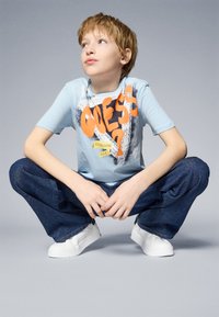 T-shirt de color azul claro con texto gráfico naranja y un diseño de salpicaduras negras, combinado con unos pantalones vaqueros de pierna ancha azul oscuro y zapatillas blancas.