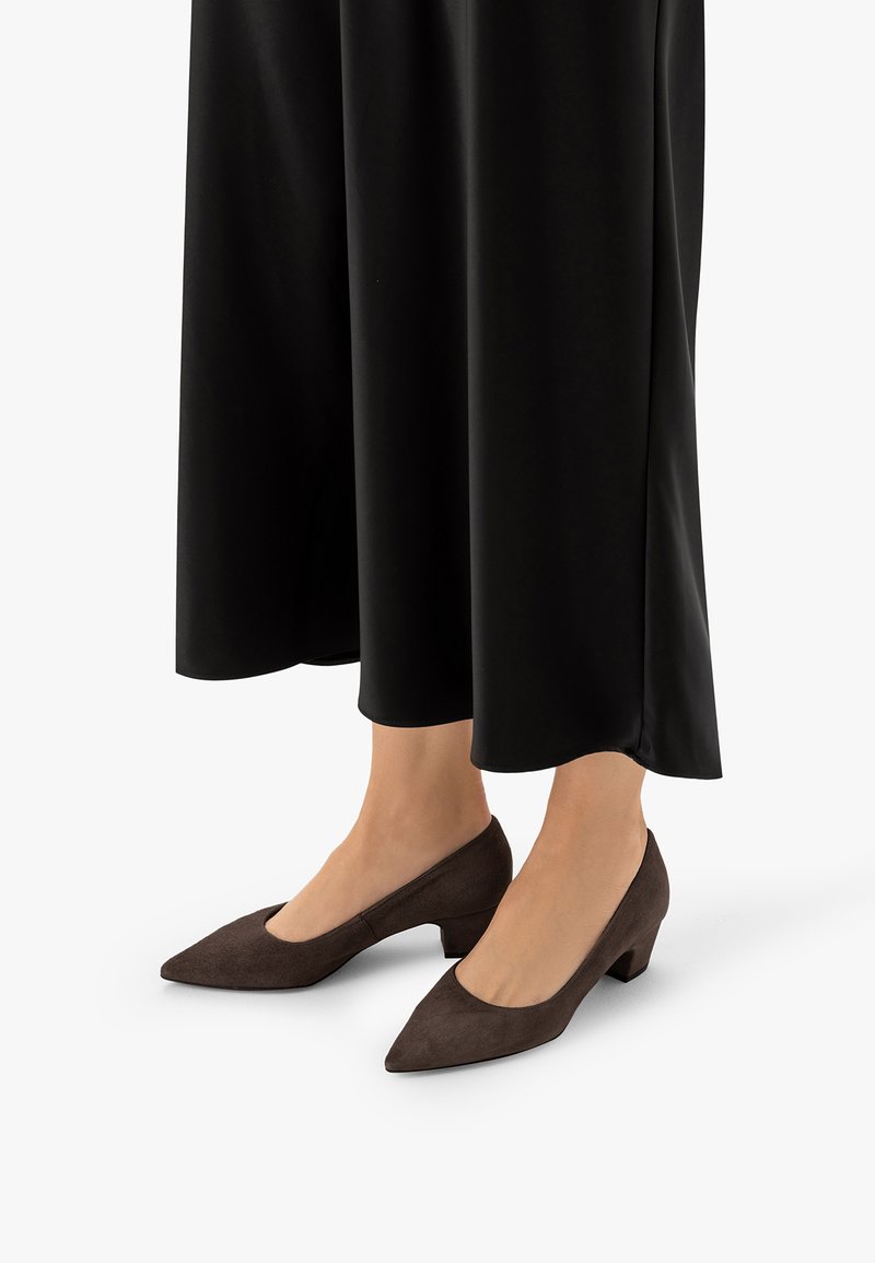 Jambes vêtues d’un pantalon noir à jambes larges et de chaussures pointues à talons bas marron foncé sur fond blanc.