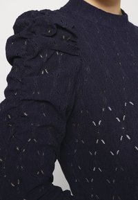 Námořnický pulovr s texturovanou tkaninou a otevřeným pletením, s volánovými rukávy. Vysoký límec a přiléhavý střih s jemnými detaily.