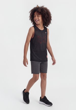 Garçon aux cheveux bouclés souriant, marchant, portant un débardeur en mesh noir Nike, un short gris foncé et des baskets noires.