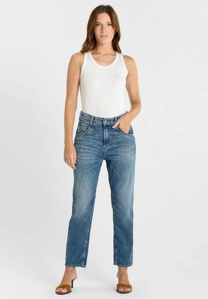 Vrouw staand, gekleed in een witte mouwloze top, blauwe jeans met hoge taille en rechte pijpen, en bruine open-toe sandalen met hakken, tegen een effen witte achtergrond.