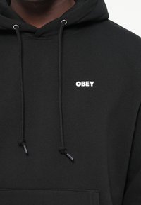 Hanorac negru din bumbac moale, cu un buzunar frontal, având un logo alb "OBEY" pe pieptul stâng și șnururi ajustabile.