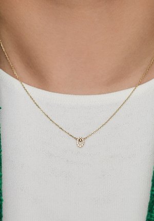 Collier chaîne dorée avec un petit pendentif ovale entouré de pierres précieuses claires, porté sur un haut blanc.