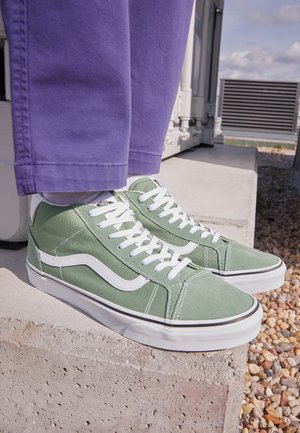 Personne portant des baskets montantes Vans vertes avec des lacets blancs et un pantalon violet debout sur des marches en béton à l'extérieur.