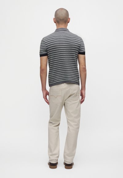 Homme debout de dos portant une chemise à manches courtes rayée grise et blanche, un pantalon beige clair et des chaussures marron sur un fond uni.