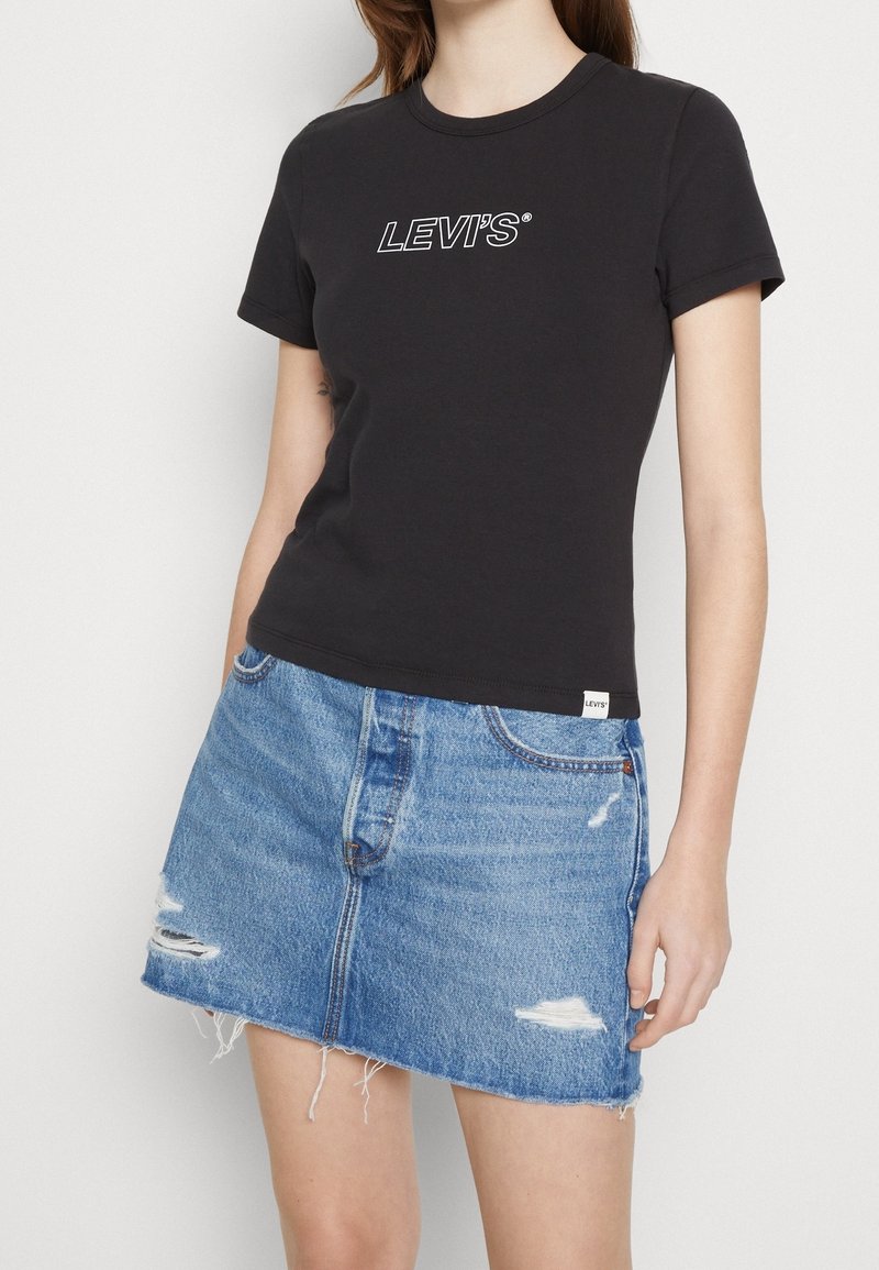 T-shirt en coton noir avec le logo « LEVI’S » blanc, associé à une jupe en denim bleu avec des détails déchirés et un ourlet effiloché.