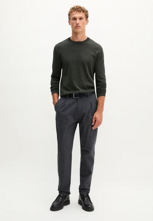 Homme debout, mains dans les poches, portant une chemise longue sombre, un pantalon gris avec ceinture et des chaussures en cuir noires sur un fond uni.