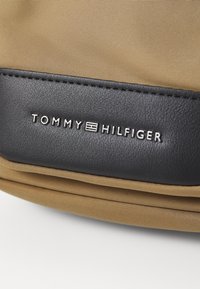 Geantă din nylon bej cu un accent din piele neagră. Prezintă logo-ul „TOMMY HILFIGER” embosat. Textură netedă cu un design structurat și cusături.