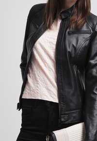 Chaqueta de motociclista de cuero negro con un diseño ajustado, bolsillos con cremallera y una parte superior acolchada de color rosa claro debajo. Jeans oscuros y un bolso de crema.