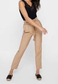 Pantaloni beige a vita alta con cintura legata. Tessuto liscio, gamba affusolata, parte anteriore con pieghe, abbinati a scarpe nere slip-on e un top in pizzo nero.