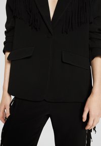 Černý blazer s třásněmi podél límce, dvěma předními kapsami a odpovídajícími černými kalhotami s třásněmi po stranách.