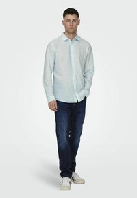 Lichtblauw, lange mouwen blouse met knopen, van een katoenmix, gedragen met donkere spijkerbroeken en witte sneakers, staande pose tegen een effen achtergrond.