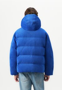 Person set fra ryggen iført en blå kordfløjl pufferjakke med hætte og lyseblå denim jeans mod en ensfarvet baggrund.