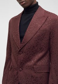 Homme portant un blazer rouge foncé à motifs par-dessus un pull à col roulé noir, visible du torse au cou.
