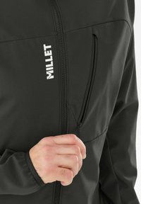 Veste noire avec une poche poitrine zippée et des poignets élastiques, fabriquée en matériau doux et extensible ; comporte un logo "MILLET" blanc.