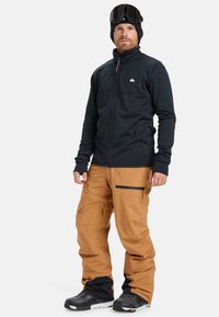 Schwarze Fleecejacke mit Reißverschluss, beige Schneehose mit Taschen und schwarze Schneestiefel; Träger trägt eine Skibrille und eine Mütze.