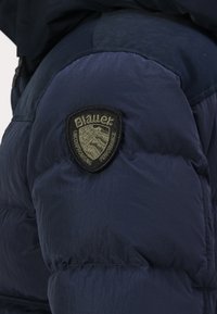 Mornarsko modra podložena jakna z teksturirano površino in črnim logotipom na rokavu, embosiranim z "Blauer" in "Uncompromising Performance."