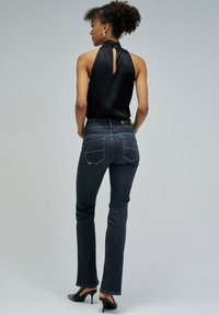 Mujer de espaldas con una blusa sin mangas negra, jeans oscuros ajustados, zapatos negros de tacón alto y pendientes plateados sobre un fondo gris liso.