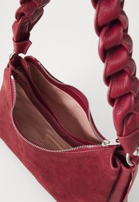 Red suede handbag with a braided strap, unzipped to reveal a spacious interior lined in light fabric and featuring a brand label.