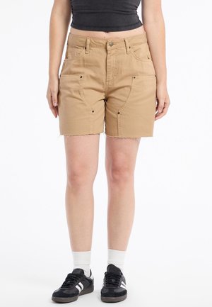 Shorts en coton de couleur tan, arrivant au genou, avec poches avant et coutures latérales. Détail d'ourlet effiloché. Associés à des baskets noires et des chaussettes blanches.