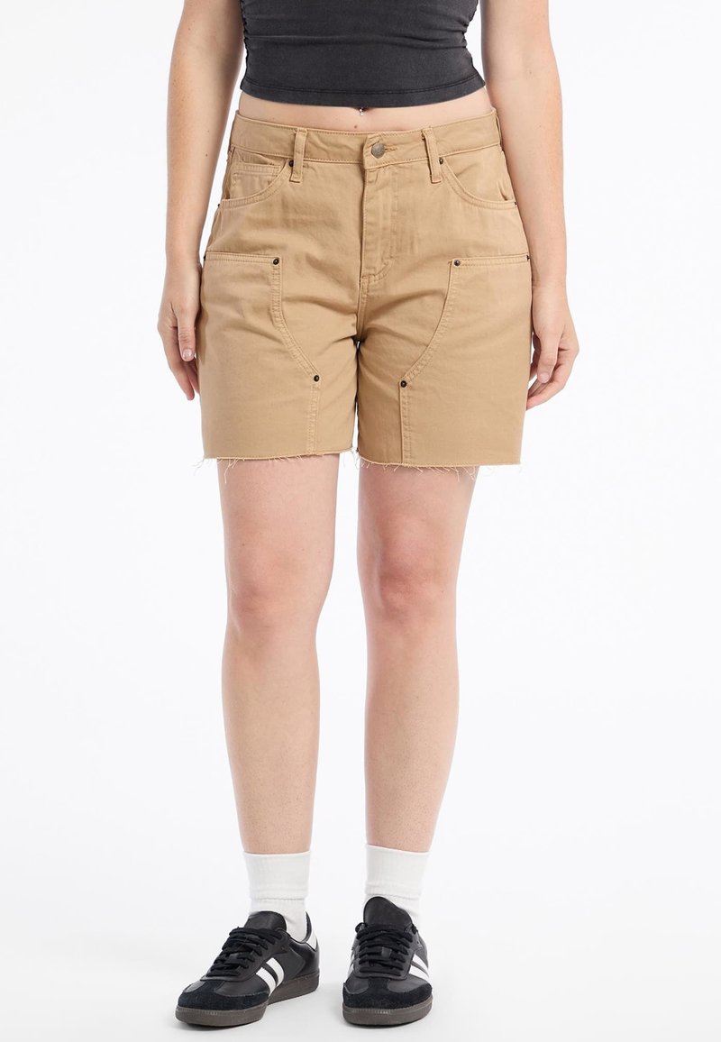 Pantaloni corti color sabbia, fino al ginocchio, in cotone, con tasche anteriori e cuciture laterali. Dettaglio di orlo sfilacciato. Abbinati a sneaker nere e calzini bianchi.