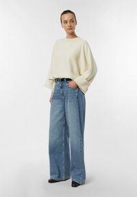 Cream knit sweater with wide sleeves paired with high-waisted light blue wide-leg jeans. Black belt with round buckle, casual footwear.