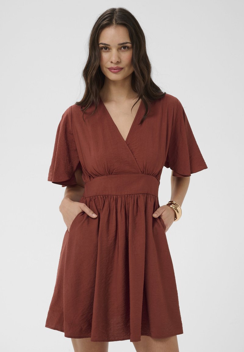Femme aux longs cheveux noirs portant une robe couleur rouille à encolure en V et manches courtes, posant les mains dans les poches devant un fond uni.