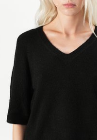 Svart strikket genser med V-hals, korte ermer og en myk tekstur. Stoffet ser varmt og koselig ut med en litt løs passform.