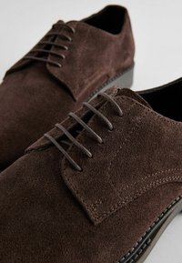 Zapatos de ante marrón con puntera redondeada, seis ojales para los cordones y una costura decorativa a lo largo del lateral y la suela.