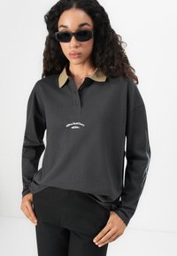 Kvinne med langt krøllete hår iført svarte solbriller, en mørkegrå langermet polo med Quiksilver-logo, og svarte bukser, som står med hendene på hoftene.