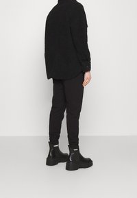 Svart fleece-tröja med långa ärmar, avslappnad passform och knäppning vid ärmsluten, i kombination med svarta tapered joggers och kraftiga svarta ankelstövlar.