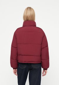 Chaqueta de plumas burdeos con cuello alto, diseño acolchado y puños elásticos, que presenta una silueta corta y una textura suave.