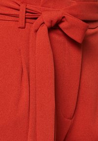 Pantalons en tissu rouge avec une large ceinture à nouer et une poche latérale, affichant une surface lisse et texturée.