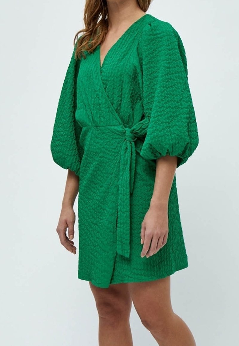 Femme portant une robe portefeuille verte texturée avec des manches trois-quarts bouffantes et une ceinture nouée à la taille, debout devant un fond clair uni.