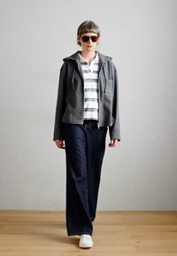 Sivá mikina na zips s prúžkovanou polokošeľou, široké tmavé džínsy a biele tenisky. Doplnky zahŕňajú hnedé slnečné okuliare.